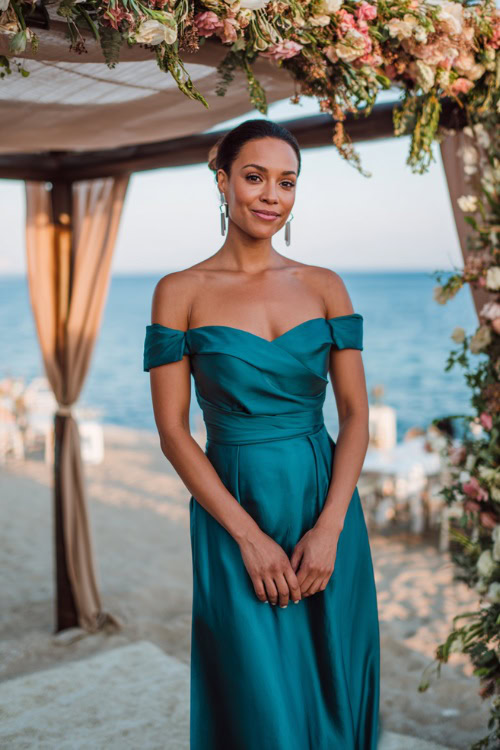 A woman wearing a teal off-shoulder gown with a subtle train and delicate earrings, standing near a floral-covered pergola at a destination wedding by the sea