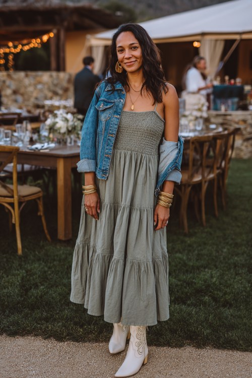 A woman wears a sage green tiered boho maxi dress with white cowboy boots and a denim jacket, accessorized with gold bracelets