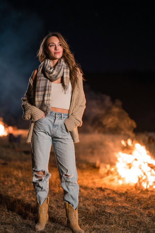 A woman wears light wash ripped jeans with a cozy oversized cardigan, styled with cowboy boots and a plaid scarf, standing by a bonfire at a country concert under fall night skies