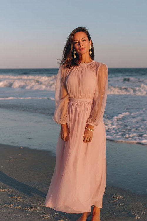A woman wears a blush pink chiffon maxi dress with long sheer sleeves, paired with delicate sandals and pearl earrings
