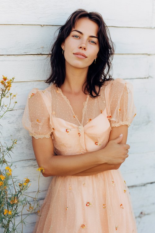 A woman wears a pastel peach dress with tiny embroidered blooms and short sheer sleeve