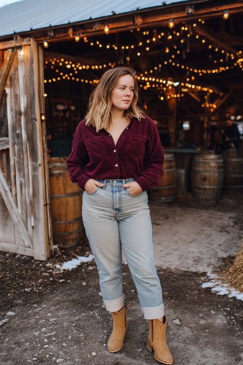 A curvy woman wears light-wash straight-leg jeans cuffed at the ankle, a burgundy button-up flannel, and tan short cowboy boots
