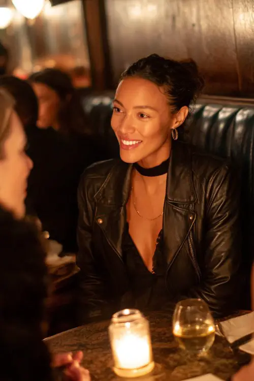 A woman over 40 wears a black midi dress with a high neckline and leather jacket, chatting with friends at a candle-lit booth
