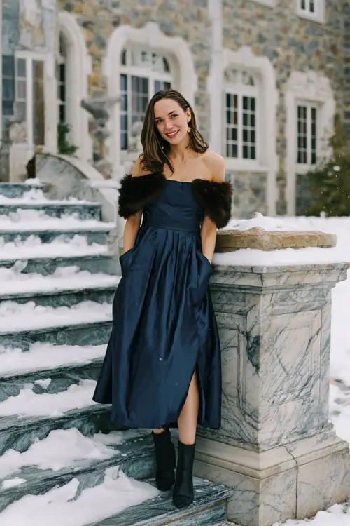 A woman over 40 wears a navy blue off-the-shoulder gown with a pleated skirt and fitted bodice, paired with black heeled boots