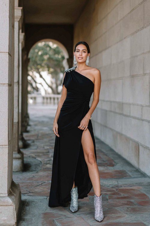 A woman wears a black one-shoulder crepe gown with a thigh slit and a silver capelet, styled with silver glitter ankle boots and statement earrings