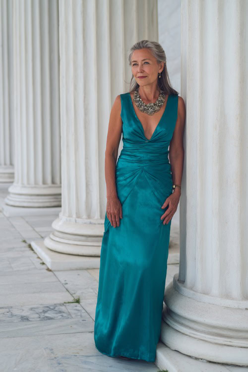 A woman over 50 in a teal satin evening dress with a wide V-neckline and statement necklace, posing near a white columned building during a formal wedding