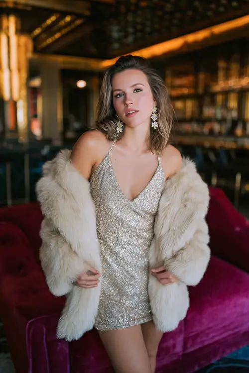 A woman wears a glittery halter neck mini dress with an oversized faux fur jacket and chandelier earrings