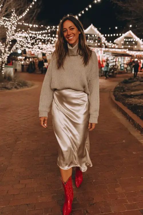 A woman wears a champagne satin midi skirt with a taupe mock-neck sweater and bold red cowboy boots