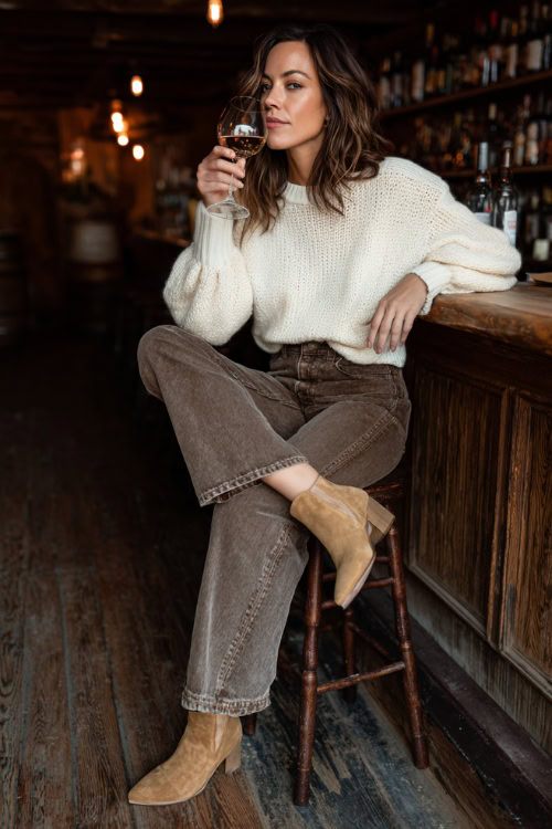 A woman in wide-leg jeans, a fitted cream sweater, and suede ankle boots, sipping wine at a casual fall bar with wooden interiors, full body outfit visible