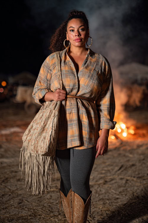 A curvy woman wears a belted plaid shacket over leggings, styled with cowboy boots and hoop earrings, holding a fringed bag at a fall concert bonfire setting