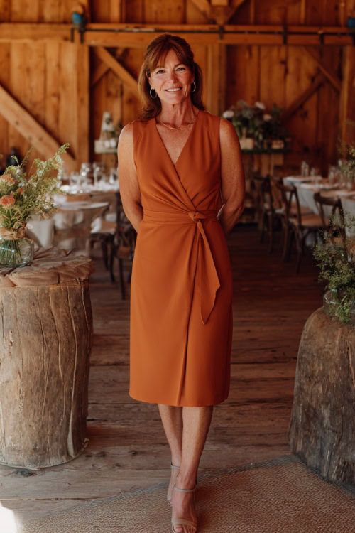 A woman over 50 in a terracotta-colored sleeveless wrap midi dress with minimal jewelry and neutral sandals, standing near rustic wooden deÌcor at a barn-inspired cocktail wedding