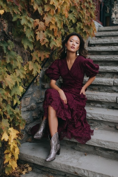 A woman over 30 wears a burgundy lace high-low dress with puffed sleeves, styled with metallic cowboy boots and pearl drop earrings