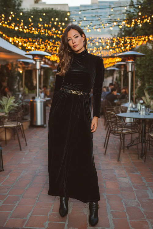 A woman wears a black velvet long-sleeve gown with a high neckline, accessorized with a metallic belt and heeled boots
