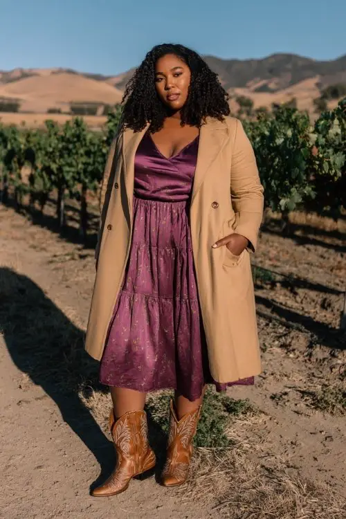A curvy woman in a plum satin A-line dress and a layered skirt, paired with brown embroidered cowboy boots and a lightweight camel coat