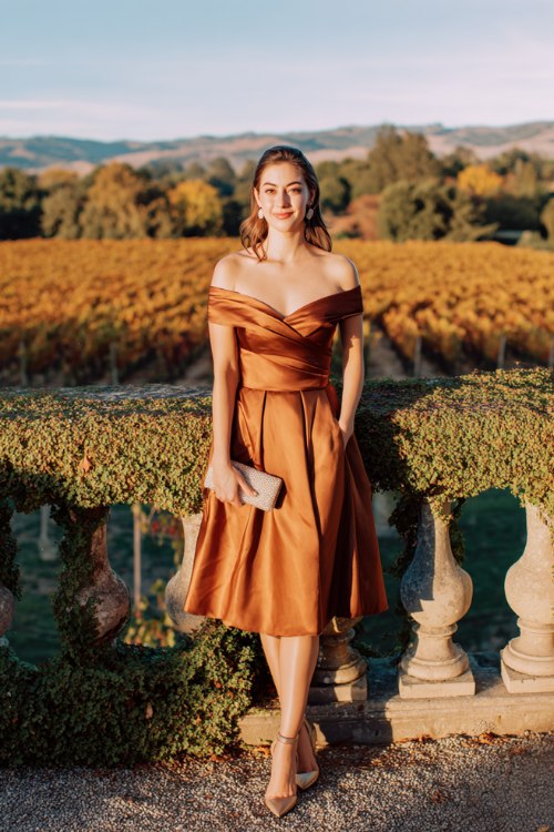 A petite woman wears a copper satin cocktail dress with pleated skirt and off-shoulder neckline, paired with metallic gold heels