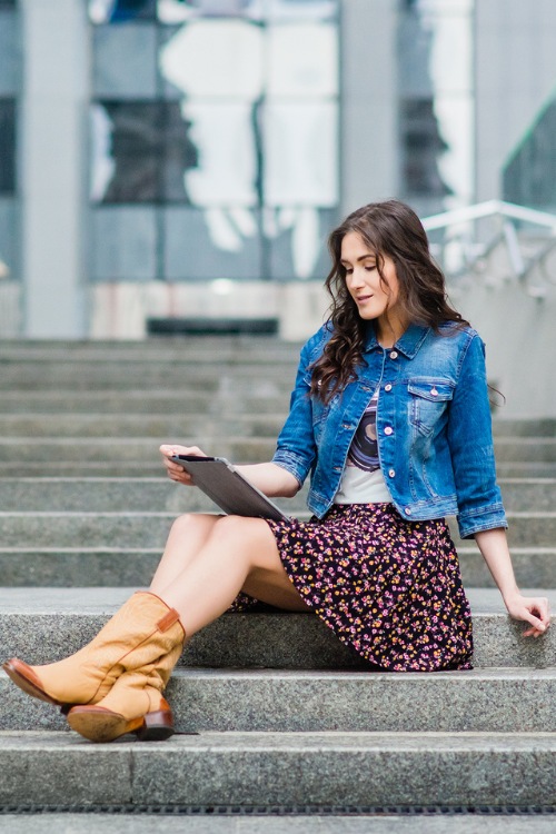 A woman wears brown cowboy boots with a floral dress and a denim coat