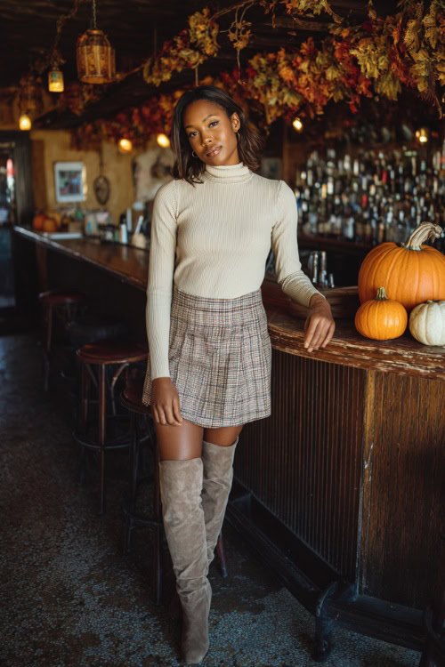 A Black woman wearing a plaid mini skirt, a fitted cream turtleneck, and over-the-knee suede boots, standing near a cozy bar with warm lights and pumpkins at the entrance