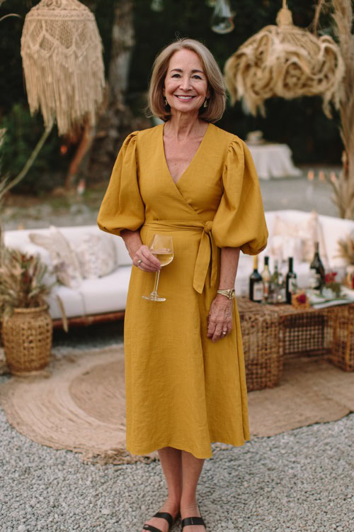 A woman over 50 wearing a mustard yellow belted midi dress with subtle puff sleeves and espadrille wedges, smiling near a boho-chic cocktail wedding setup
