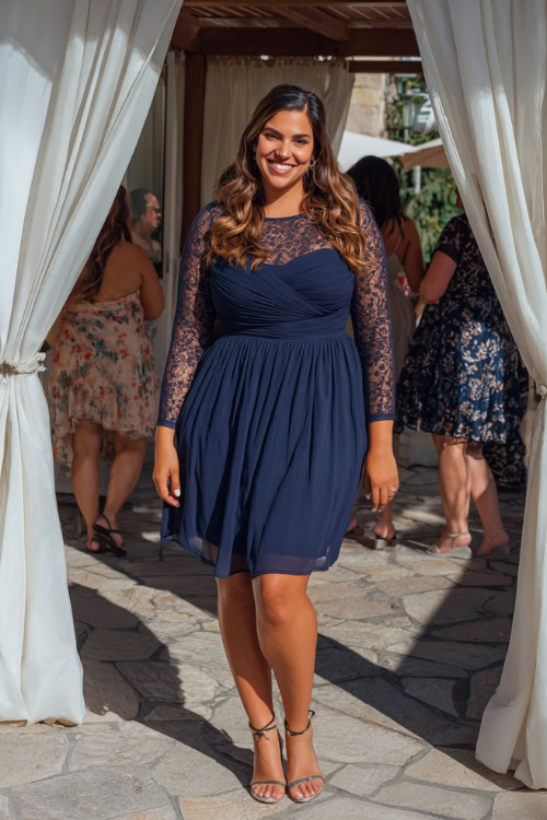 A plus-size woman in a navy chiffon midi dress with lace sleeves and low heels, standing under a white draped pergola at a summer wedding reception
