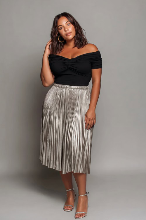 A plus-size woman wearing a fitted black off-shoulder top with a pleated midi skirt and metallic sandals, styled for a casually elegant summer going-out outfit