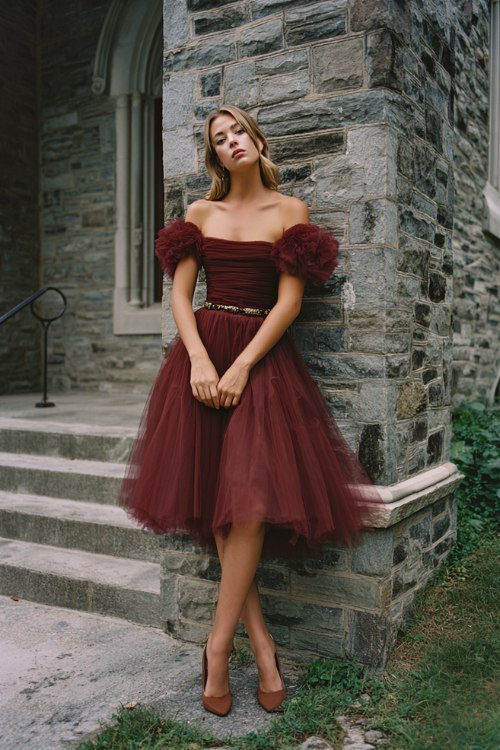 A woman wears a burgundy off-shoulder cocktail dress with tulle layers and a crochet bodice, finished with suede block heels