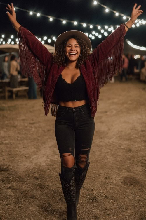 A curvy woman wears ripped black jeans with tall black cowboy boots and a burgundy fringe jacket