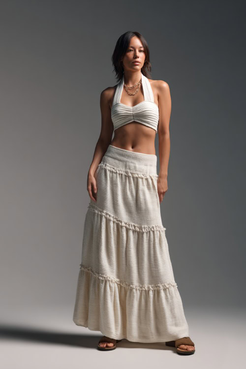 A woman wearing a knit halter top with a tiered maxi skirt and flat sandals, styled for a laid-back summer evening, simple studio background