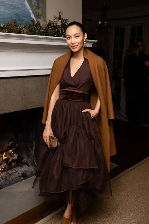A woman wears a chocolate-brown organza gown with layered skirt and velvet waistband, layered with a camel wool cape coat and suede ankle-strap heels