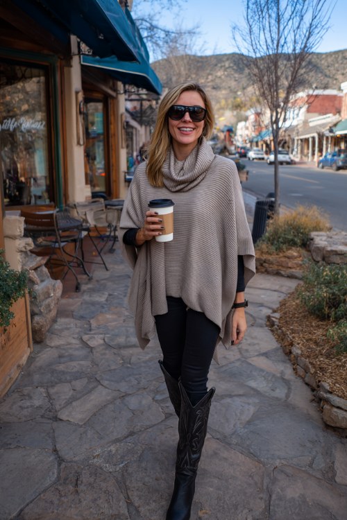 A woman over 40 wears black leggings with sleek tall cowboy boots and a soft cashmere poncho, holding a coffee cup while strolling through a cozy town street