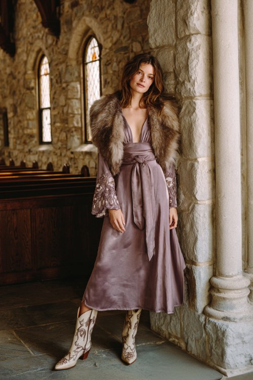 A woman wears a long-sleeve mauve dress with sheer embroidered sleeves and a satin sash, layered with a faux fur capelet in dove grey and taupe floral cowboy boots