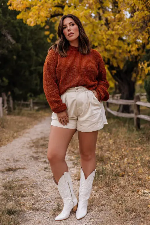 A plus size woman wears white cowboy boots with khaki shorts and a rust-colored ribbed sweater