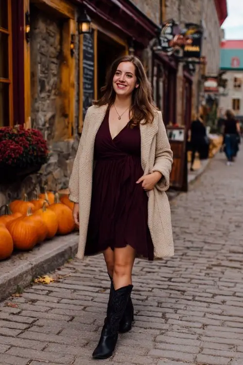 A curvy woman wears a burgundy wrap dress with tall black cowboy boots and a beige wool coat