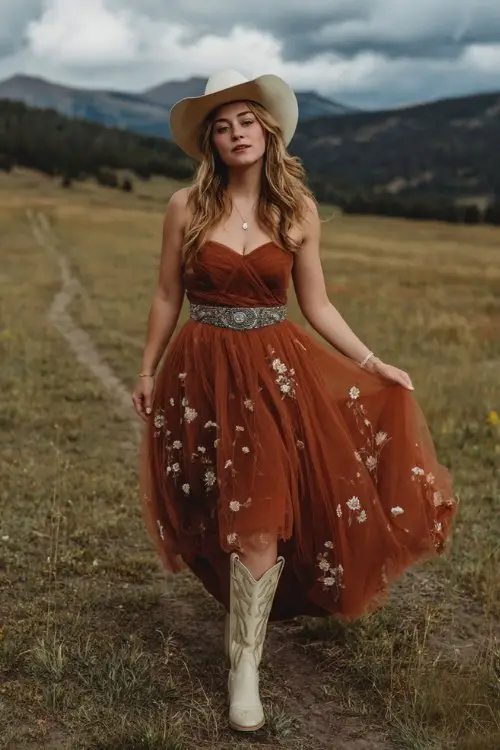 A curvy woman wears a rust tulle gown with embroidered wildflowers across the skirt, paired with cream cowboy boots and a concho belt (2)