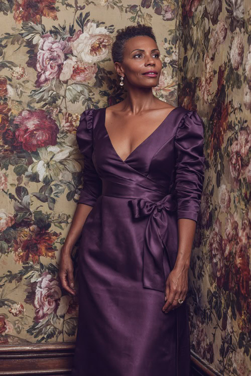 A woman over 50 in a deep purple formal dress with three-quarter sleeves and satin sash, posing beside a floral wall backdrop at a luxurious wedding reception
