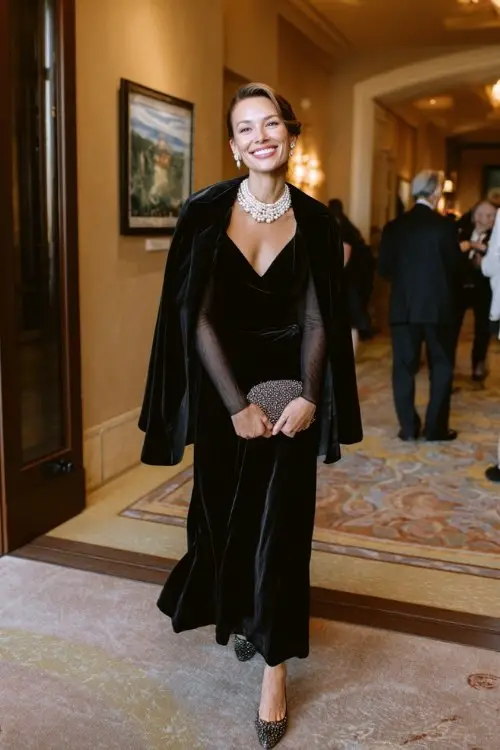 A woman wears a black velvet A-line dress with long mesh sleeves, paired with a pearl-embellished wool cape and sparkly pumps