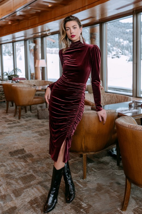 A woman wears a wine-colored high-neck velvet dress with side ruching and patent leather boots