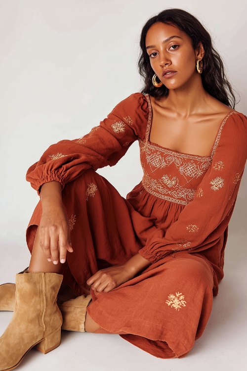 A woman wearing a rust-colored midi dress with tonal floral embroidery, paired with tan suede ankle boots and gold hoop earrings