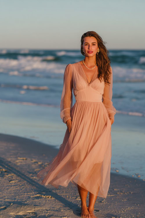 A woman wears a blush pink chiffon maxi dress with long sheer sleeves, paired with delicate sandals and pearl earrings