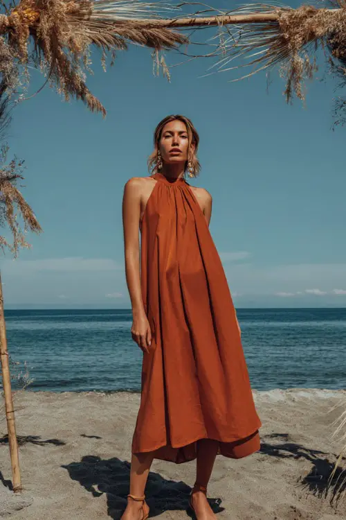 A woman wears a rust-orange sleeveless midi dress with a pleated skirt, paired with block heel sandals and minimal jewelry