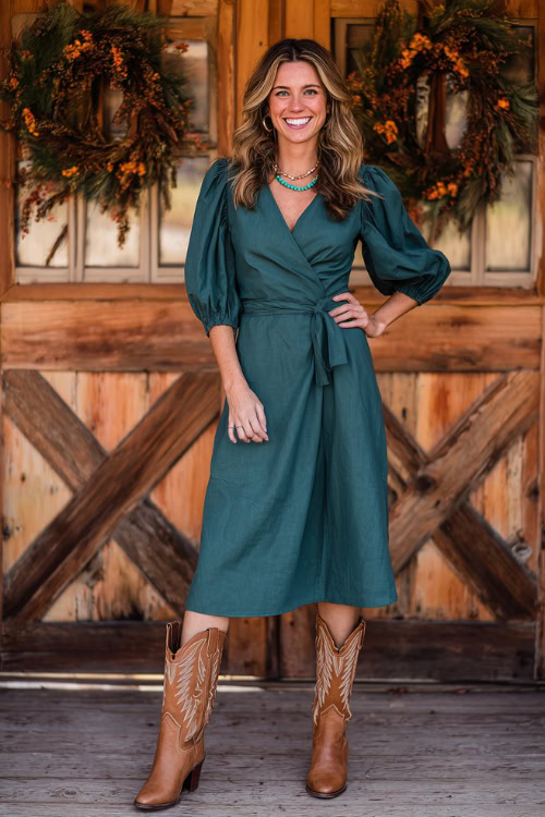 A woman wears a forest green wrap dress with puff sleeves and a belted waist, styled with tan cowboy boots and turquoise jewelry, posing in front of wooden ranch doors with fall wreaths