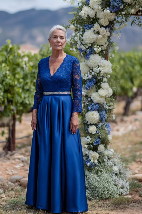 A woman over 50 wearing a royal blue A-line gown with lace sleeves and a silver belt, standing beside a floral arch in a vineyard setting, full body view