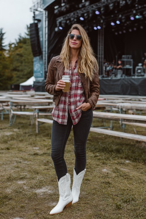 A woman over 30 wears black skinny jeans tucked into white cowboy boots with a plaid flannel shirt, layered with a leather moto jacket