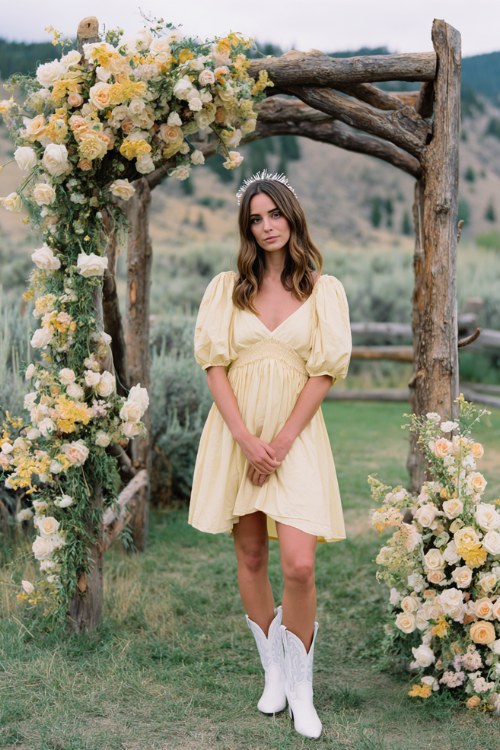 A woman wears a flowy pastel yellow dress with white cowboy boots and a delicate headband