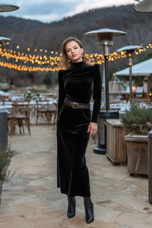A woman wears a black velvet long-sleeve gown with a high neckline, accessorized with a metallic belt and heeled boots