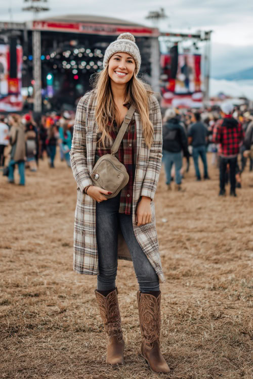 A woman wears a long plaid coat with skinny jeans, suede cowboy boots, and a crossbody bag, styled with a knit beanie at an outdoor music festival stage