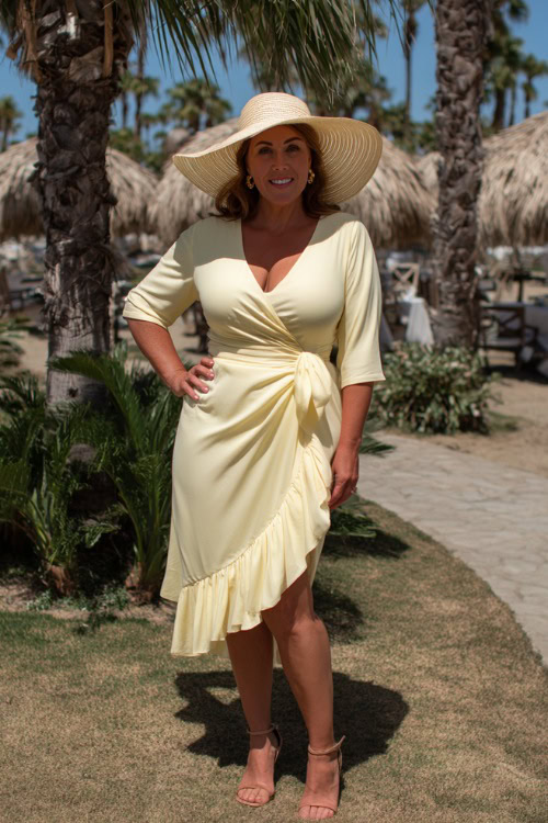 A plus-size woman over 50 in a pastel yellow wrap midi dress with ruffled hem and a wide-brim hat, posing under palm trees at a beach wedding