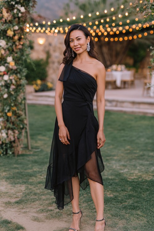 A woman wears a black one-shoulder midi dress with an asymmetrical layered hem and sheer overlay