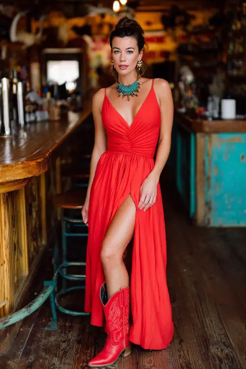 A woman wears a red V-neck maxi dress with a high slit and ruched waist, styled with turquoise statement jewelry and bright red cowboy boots