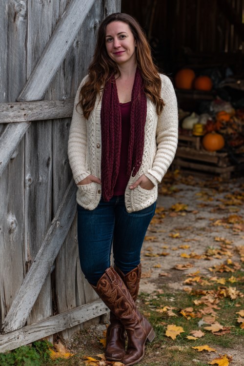 A curvy woman wears tall brown cowboy boots with dark blue skinny jeans, a cream cardigan, and a burgundy scarf