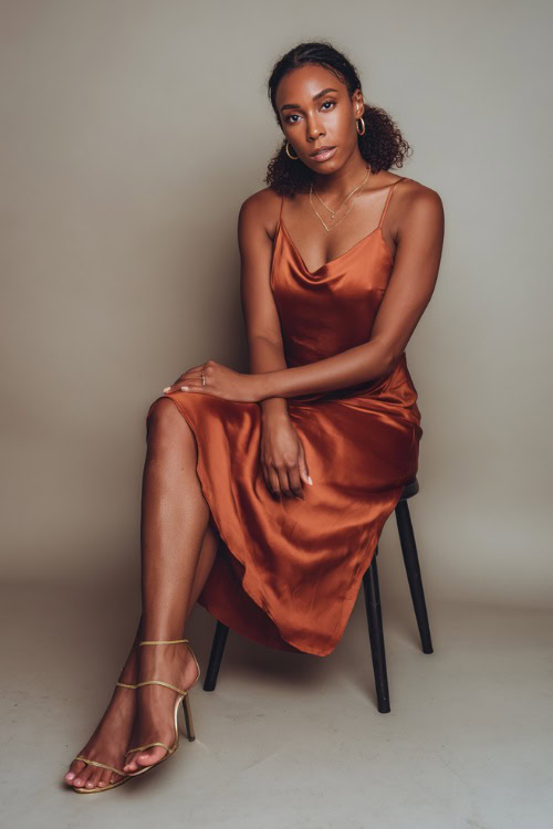 A Black woman wearing a satin rust-colored midi slip dress with gold accessories and strappy heels, styled for a classy summer dinner look, simple studio background